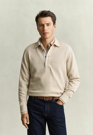 Young man with dark hair wears a beige long-sleeve polo shirt over a checked shirt, dark jeans, and a brown belt, standing with one hand in pocket.