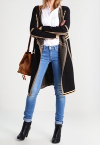 Schwarze Strickjacke mit tanfarbenen Akzenten, weißes Hemd, blaue enge Jeans und schwarze Absatzstiefeletten. Braune Tasche über der Schulter getragen.