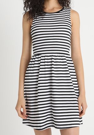 Femme portant une robe sans manches à rayures horizontales noires et blanches avec une taille froncée, se tenant devant un fond clair uni.