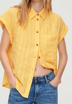 Woman wearing a yellow textured short-sleeve button-up shirt with a pocket, paired with blue denim jeans, hands in pockets.