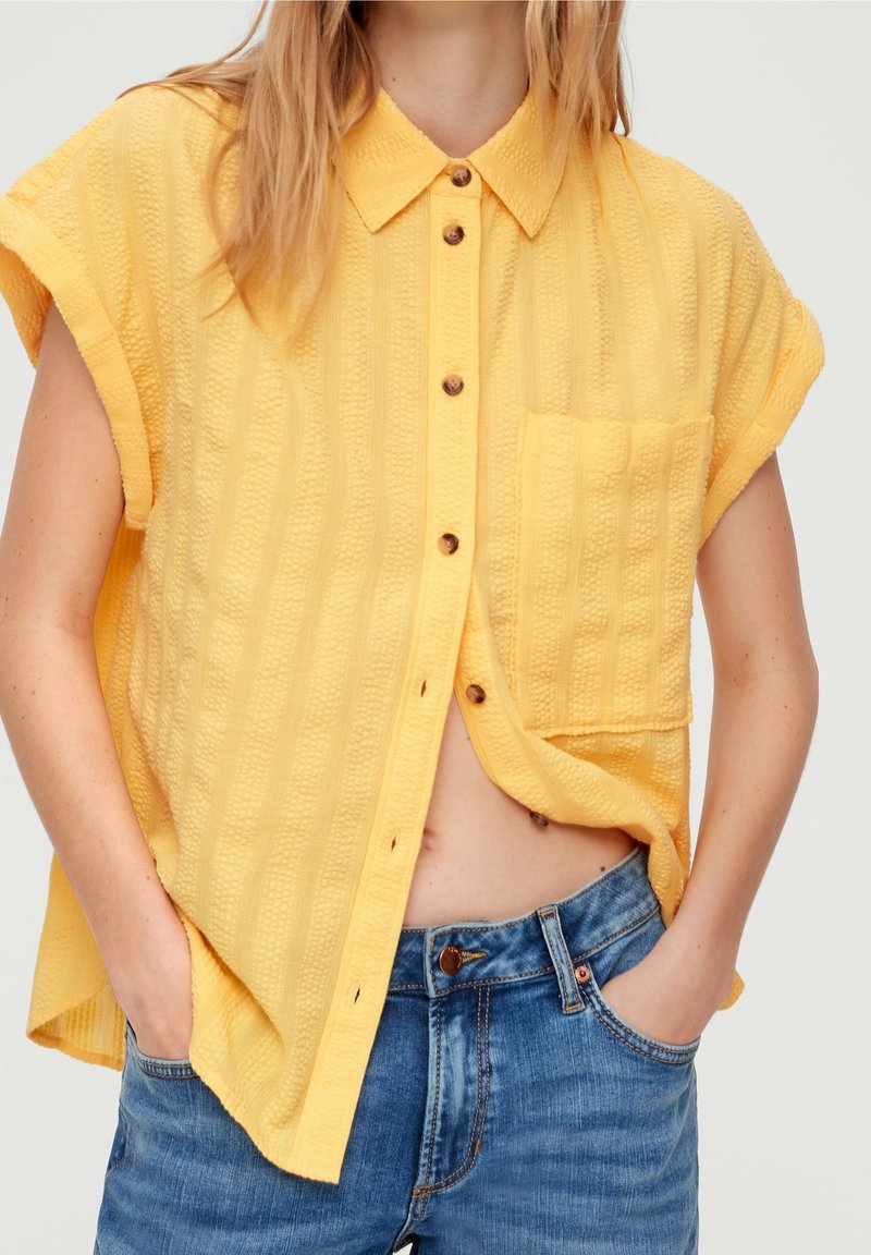 Femme portant une chemise jaune texturée à manches courtes avec une poche, associée à un jean en denim bleu, mains dans les poches.