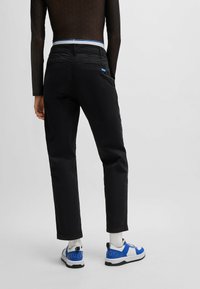 Pantalons noirs à coupe décontractée, avec poches latérales, et un patch logo bleu visible à l'arrière. Associés à des chaussures de sport blanches et bleues.