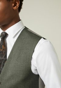 Chaleco gris texturizado con un diseño de cuello en V, usado sobre una camisa blanca. Presenta una corbata con un patrón en tonos oscuros y naranjas.