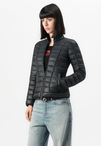 Donna che indossa una giacca trapuntata nera sopra una maglietta nera con logo rosso, abbinata a jeans azzurri chiari, in piedi con una mano in tasca.