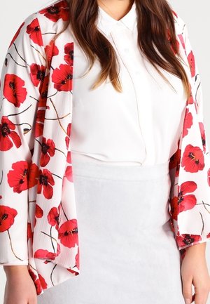 Femme portant un chemisier blanc et une jupe associée à une veste fleurie blanche avec des fleurs de pavot rouges.