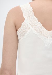 Ώμος και άνω μέρος της πλάτης ενός ατόμου που φοράει λευκό ριμπ ζιβάγκο με φαρδιές λουλουδάτες δαντελένιες τιράντες και περιθώριο.