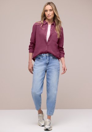 Vrouw die een mauve overhemd draagt over een wit topje, lichtblauwe jeans met hoge taille en zilveren sneakers, staand tegen een effen achtergrond.