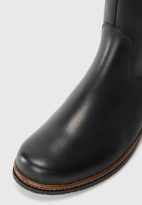 Botte noire en cuir jusqu'à la cheville avec un bout arrondi, texture lisse, semelle marron contrastante et un minimum de détails de couture le long du bord supérieur.