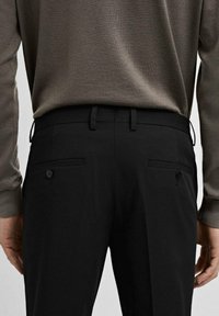 Pantalons tailleur noirs en tissu lisse, dotés de deux poches arrière, dont une avec un bouton, et un design ajusté.