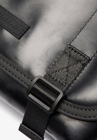 Dettaglio di una borsa in tessuto nero con texture, dotata di una fibbia in plastica nera e cuciture resistenti, arricchita da una patch rettangolare con logo.