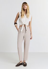 Giovane donna che indossa una camicia bianca senza maniche legata, pantaloni beige chiari con coulisse, scarpe basse scure e due braccialetti, in piedi su uno sfondo bianco.