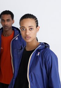 Two individuals wear lightweight jackets. The front person has a navy blue jacket with a colored zipper and logo, over a black top.