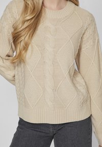 Pull en tricot beige avec un motif de torsades texturé, un col et un ourlet côtelés. Associé à un jean gris foncé, mettant en valeur le contraste des couleurs.