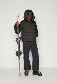 Muž v černé lyžařské bundě, černých lyžařských kalhotách, botách a brýlích drží pár lyží, stojící na jednobarevném pozadí.