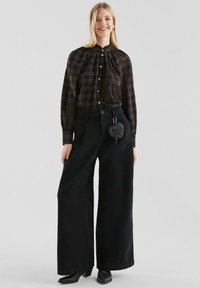 Geruite bruine blouse met een hoge halslijn, knoopsluiting en brede zwarte pantalon. Kleine gestructureerde zwarte hartvormige tas bevestigd aan de taille.