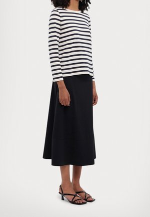 Osoba ubrana w pasiasty top z długim rękawem, czarną spódnicę midi i czarne sandały na obcasie, stojąca na tle jasnej, gładkiej ściany.