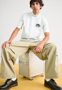 Γκρι βαμβακερό t-shirt κοντό μανίκι με κεντημένο λογότυπο, σε συνδυασμό με μπεζ παντελόνι φαρδιάς παντελούς και μαύρα παπούτσια, καθισμένο σε μια ασημένια θήκη.