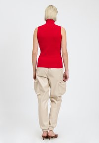 Rød ærmeløs turtleneck-top parret med beige cargo-bukser, der har store sidelommer og elastiske manchetter, sat mod en ensfarvet baggrund.