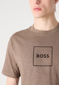 Brun bomull T-skjorte med en svart rektangulær logo med teksten "BOSS" i fet, rund skrift, samt en ribbet rund hals.