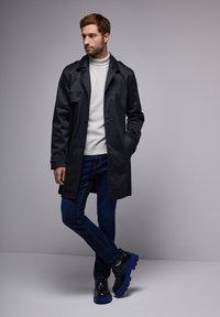 Abrigo negro con cierre de botones sobre un suéter de cuello alto gris claro, combinado con zapatos negros con detalles azules y jeans de mezclilla azules.