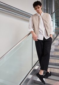 Béžová bezrukávová vesta přes bílou košili, černé zkrácené kalhoty a černé slip-on boty. Hladké tkaniny a minimalizované detaily kovových prvků.
