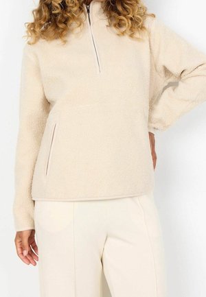 Personne portant un pull polaire crème avec une fermeture éclair à moitié et un pantalon beige clair, posant avec une main sur la hanche.