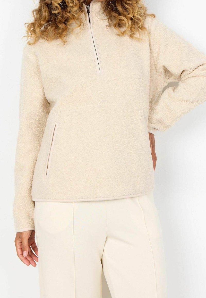 Personne portant un pull polaire crème avec une fermeture éclair à moitié et un pantalon beige clair, posant avec une main sur la hanche.