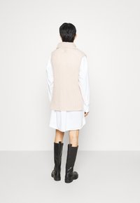 Ärmellose Strickweste in zartem Pink mit gerippter Textur und hohem Kragen, über einem weißen Hemd mit Knöpfen getragen. Schwarze kniehohe Stiefel.