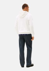 Sudadera blanca de algodón con capucha ajustable, combinada con pantalones azul oscuro. Detalle de costura visible y una camisa de capas debajo.