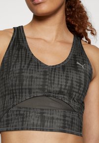 Zbliżenie na kobietę w czarnym sportowym biustonoszu z wzorem, z siateczkowym wycięciem i logo Puma na piersi.