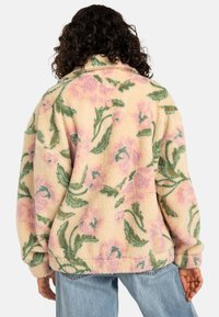 Fleecejakke med blomstermønster i pastellrosa og grønn på en kremfarget bakgrunn; har en avslappet passform og nedbrettbar krage.