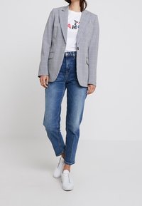 Blazer a scacchi in bianco e nero, abbinato a una maglietta grafica bianca e jeans blu. Sneakers bianche completano il look casual.