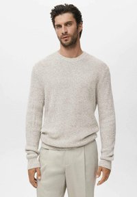 Man met kort zwart haar en een baard, gekleed in een lichtbeige gestructureerde trui en bijpassende beige broek, staande tegen een witte achtergrond.