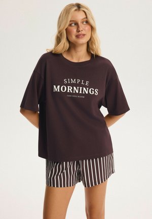 Frau mit blondem Haar, die ein dunkles übergroßes T-Shirt mit dem Schriftzug "Simple Mornings" und gestreifte Shorts trägt, steht vor einem einfarbigen hellen Hintergrund.