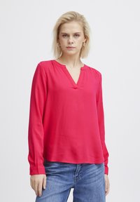 Röd långärmad blus med V-ringning, tillverkad av ett mjukt tyg. Har knäppningar vid ärmsluten och en avslappnad passform. Bärs med avslappnade blå jeans.