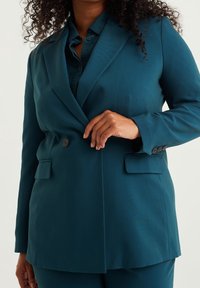 Blågrön kavaj i ett mjukt texturerat tyg med enkelknäppt fram, två framfickor och knäppbara ärmslut, kombinerad med en matchande skjorta.