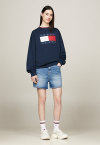 Felpa blu marino con logo rosso e bianco, abbinata a pantaloni corti in denim azzurro chiaro e sneakers bianche con dettagli rossi e neri.
