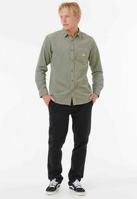 Grøn skjorte i ribfløjl med knapper foran og en brystlomme, kombineret med sorte bukser og sorte sneakers med hvide detaljer. Enkel og funktionel design.