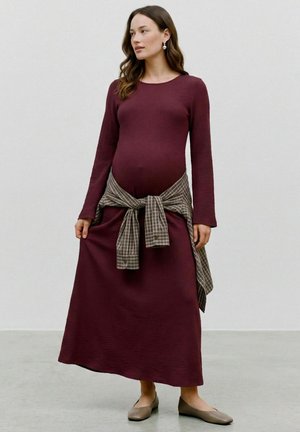 Femme enceinte portant une longue robe bordeaux avec une chemise à carreaux nouée autour de la taille et des chaussures plates beiges, debout à l'intérieur.