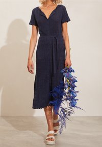 Donna che indossa un vestito midi lavorato a maglia blu navy con bottoni, mentre tiene un mazzo di fiori blu-violacei, camminando su un pavimento chiaro.