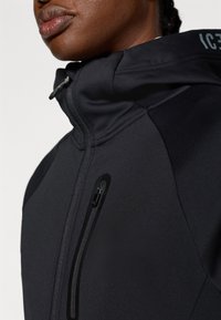 Musta tekninen takki korkealla kauluksella ja vetoketjulla edessä. Ominaisuuksiin kuuluu tyylikäs muotoilu, sileä pinta ja pieni vetoketjullinen rintatasku.