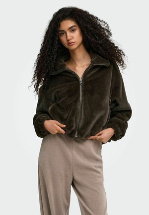 Woman with long curly hair wearing a dark brown plush zip-up jacket and light beige loose pants, standing against a plain background.