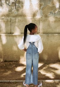 Blouse blanche légère à col volanté, associée à une salopette en denim bleu avec jambes larges et poches arrière. Chaussures décontractées visibles.