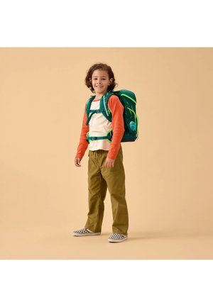 Child smiling, standing with a large green backpack, wearing an orange long-sleeve shirt, beige vest, olive pants, and checkered slip-on shoes.