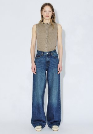 Jeune femme debout sur fond blanc, portant un haut beige sans manches boutonné et un jean bleu à jambes larges avec des baskets blanches.