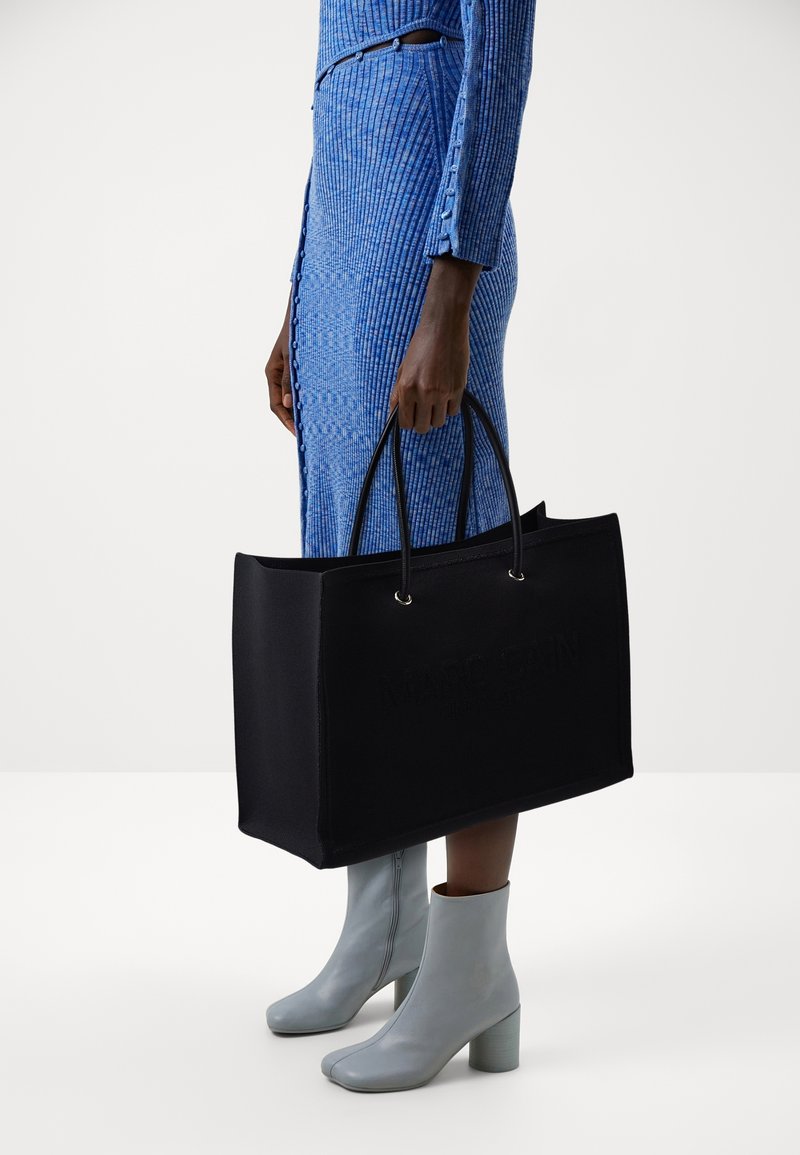 Personne en robe côtelée bleue et bottines grises tient un grand sac cabas noir par les poignées sur fond blanc.