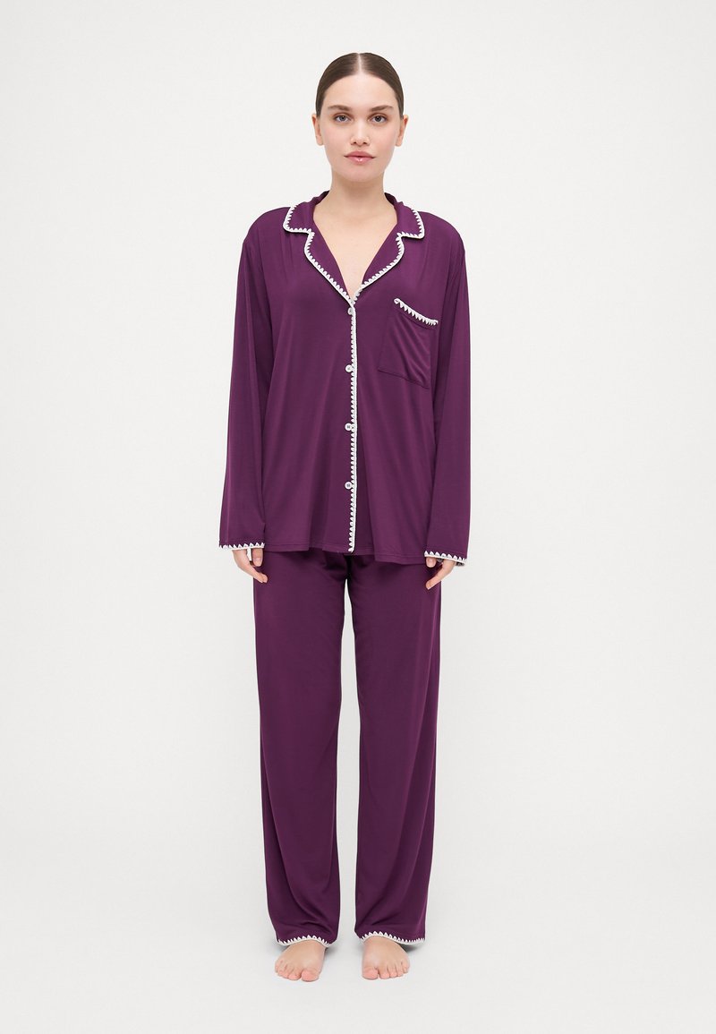Set de pijama violet, cu o cămașă cu nasturi, cu dantelă albă și un buzunar la sâni, purtată cu pantaloni asortați, largi. Textură moale.