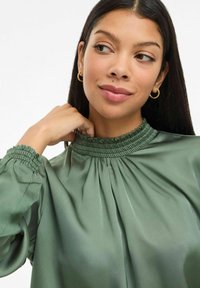 Femme aux longs cheveux bruns portant des boucles d'oreilles en or et un haut vert ruché à col montant, regardant sur le côté avec une main près du cou.