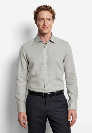 Homme aux cheveux bouclés portant une chemise gris clair boutonnée, rentrée dans un pantalon foncé avec une ceinture noire, debout devant un fond blanc uni.