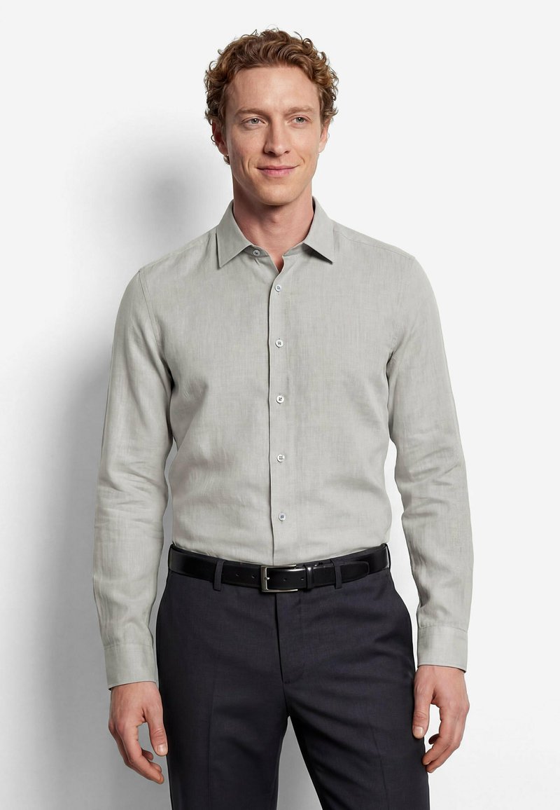 Homme aux cheveux bouclés portant une chemise gris clair boutonnée, rentrée dans un pantalon foncé avec une ceinture noire, debout devant un fond blanc uni.